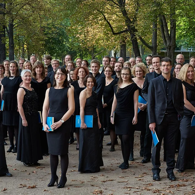 Ein großes Gruppenporträt eines Chores in formeller schwarzer Kleidung, der blaue „Bach“-Notenhefte hält und im Freien auf einem von Bäumen gesäumten Weg posiert.