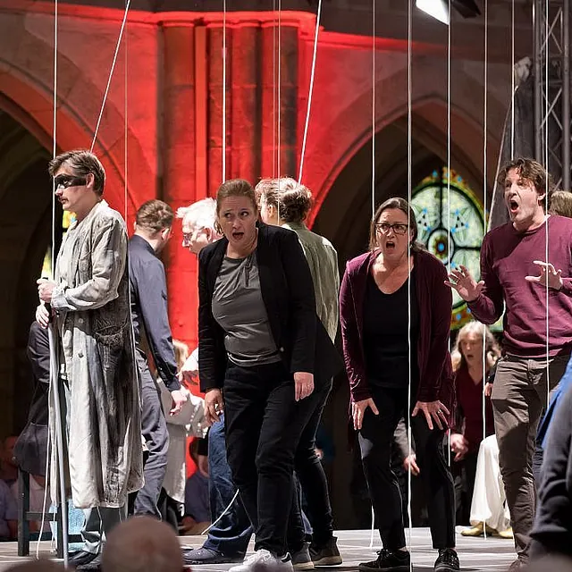 Eine dramatische Bühnenaufführung in einer Kirche, bei der mehrere Schauspieler zwischen vertikalen weißen Seilen stehen und unter rotem Licht mit intensivem Ausdruck singen.