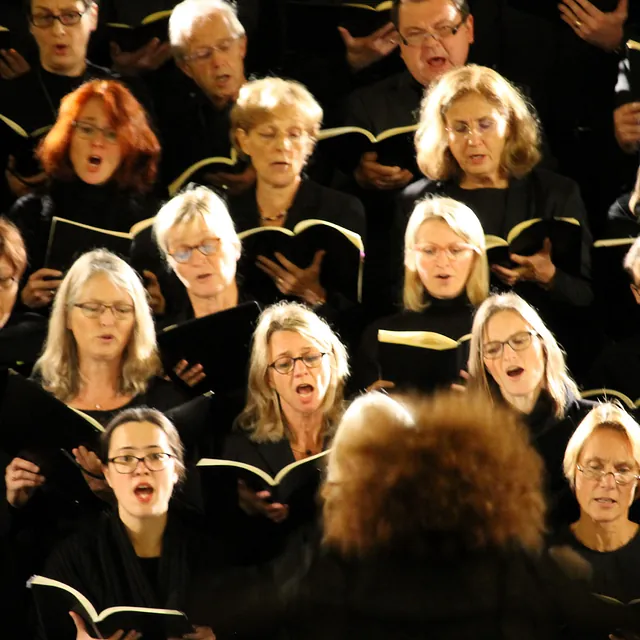 Eine Nahaufnahme von Chormitgliedern in schwarzer Kleidung, die aus offenen Notenheften singen, aus der Sicht hinter der Dirigentin.