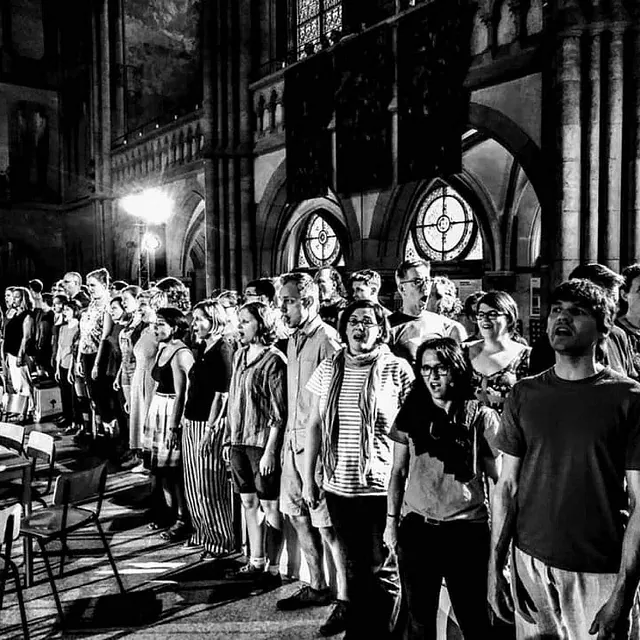 Eine kontrastreiche Schwarz-Weiß-Fotografie einer großen, vielfältigen Gruppe von Menschen, die gemeinsam in einer historischen Kirche mit hohen Deckengewölben und Buntglasfenstern singen.