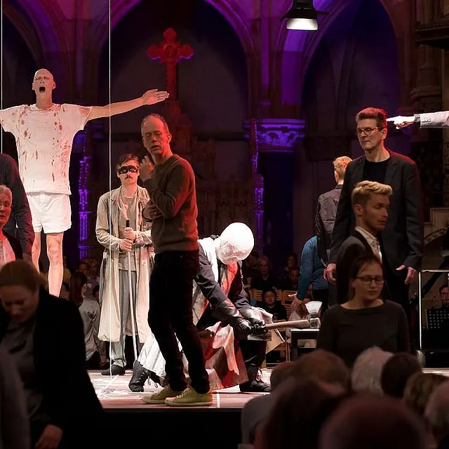 Eine belebte Bühnenszene in einer Kirche mit dramatischer Beleuchtung; zu sehen sind ein Mann mit ausgestreckten Armen in einem blutverschmierten Hemd und ein Darsteller mit goldener Krone und grauem Mantel, der über die Bühne zeigt.