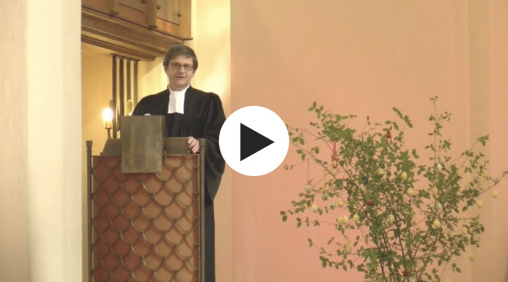 Pfarrer spricht an einer Kanzel neben einer geschmückten Pflanze.