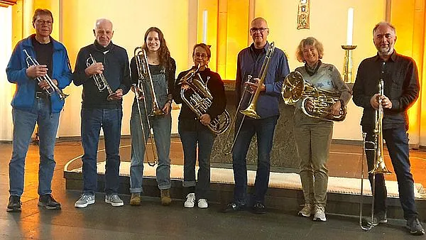 Ein Gruppenporträt von sieben Blechbläsern, die mit verschiedenen Instrumenten wie Trompeten, Posaunen und einem Euphonium vor einem Altar in einer Kirche stehen.