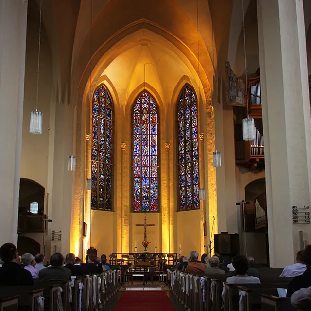 Kirchenraum mit rotem Teppich und hohen Buntglasfenstern im Altarraum.
