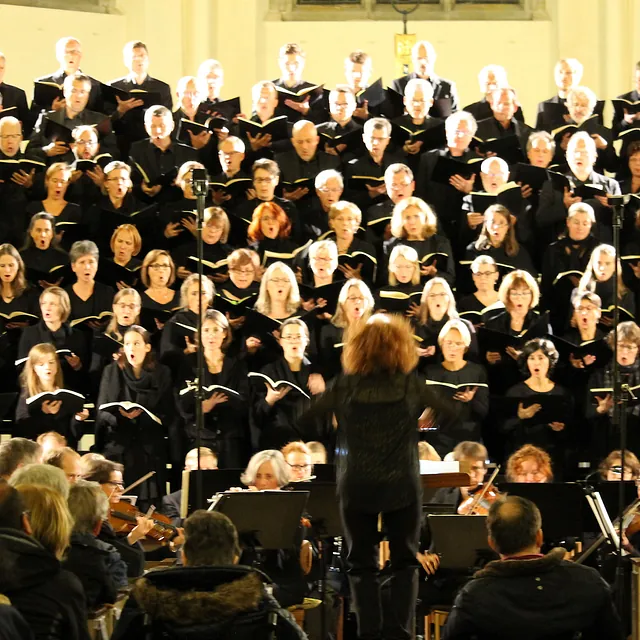 Ein großer, schwarz gekleideter Chor tritt in einer hell erleuchteten Kirche auf, begleitet von einem Orchester und geleitet von einer Dirigentin mit roten Locken.