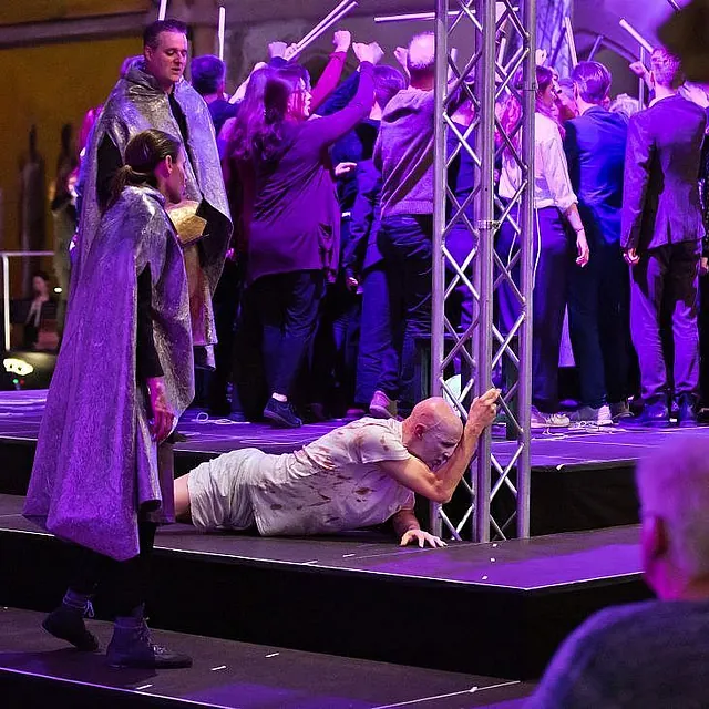 Eine dramatische Bühnenaufführung in einer Kirche mit lila Beleuchtung, bei der ein Mann in einem schmutzverschmierten Hemd auf einer gestuften Plattform auf eine Metalltraverse zukriecht, während andere Darsteller in silbernen Umhängen zusehen.