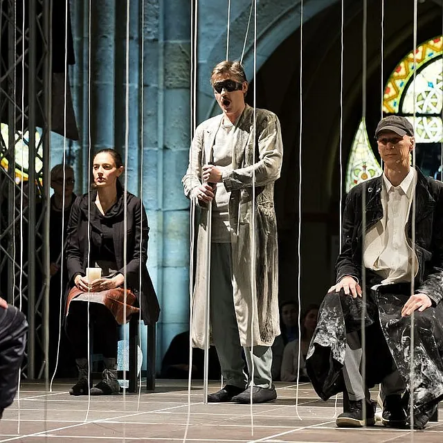 Eine dramatische Bühnenaufführung in einer Kirche, bei der drei Schauspieler zwischen zahlreichen vertikalen weißen Seilen sitzen oder stehen, die von der Decke hängen, während ein Mann mit Maske und zerschlissenem Mantel in der Mitte singt.