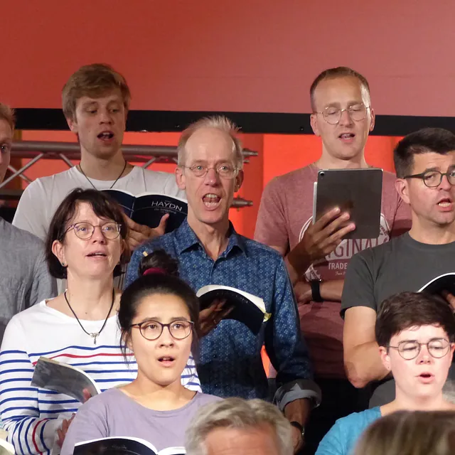 Eine Nahaufnahme von Chormitgliedern, die während einer Probe vor einem leuchtend roten Hintergrund aus Notenheften singen, darunter Haydns „Die Schöpfung“.
