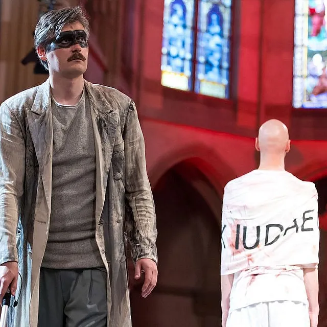 Eine dramatische Aufführungsszene in einer Kirche mit einem Mann mit Schnurrbart und einer aufgemalten schwarzen Augenmaske, der einen zerschlissenen Mantel trägt und neben einer weiteren Person steht, die dem Betrachter den Rücken zuwendet.