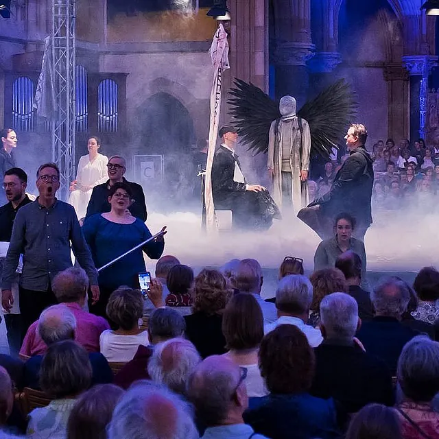 Eine dramatische Musikaufführung auf einer nebelverhangenen Bühne in einer Kirche, mit Darstellern in moderner Kleidung und einer zentralen Figur mit großen schwarzen Flügeln.