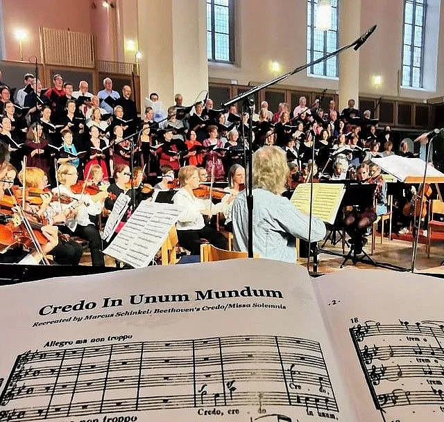 Ein Chor und ein Orchester treten in einem Kirchenraum auf, aus der Perspektive der Noten für „Credo In Unum Mundum“ von Marcus Schinkel gesehen.