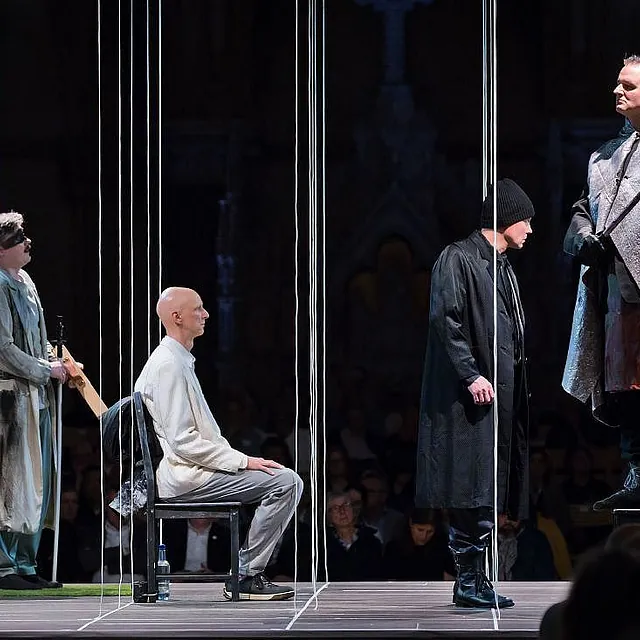 Eine dramatische Theaterszene in einer Kirche mit vier Darstellern auf einer Bühne, die durch vertikale weiße Seile unterteilt ist; ein Darsteller steht in einem schweren silbernen Umhang auf einem Stuhl, während andere in modernen Kostümen sitzen oder stehen.