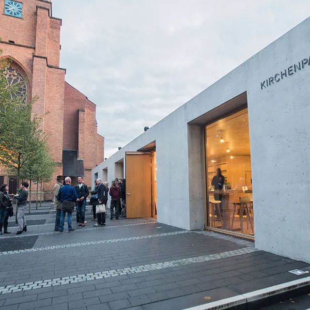 Moderner Beton-Kirchenpavillon neben einer hohen Backsteinkirche.