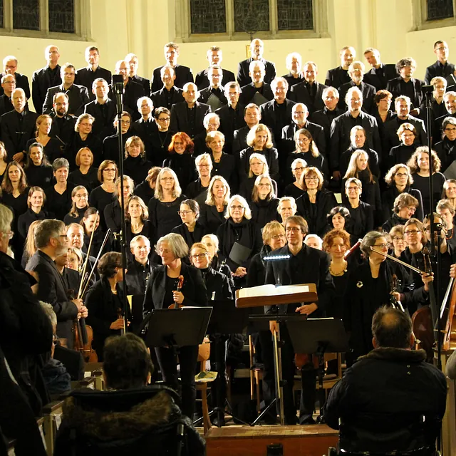 Eine Weitwinkelaufnahme eines großen, schwarz gekleideten Chores und Orchesters, die nach einem Auftritt in einem hell erleuchteten Kirchenraum stehen.