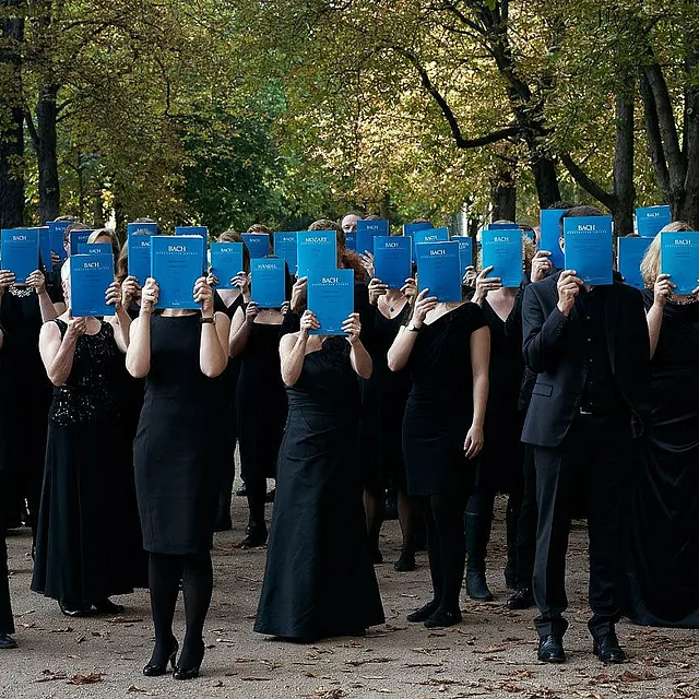Künstlerisches Gruppenporträt eines Chores in formeller schwarzer Kleidung im Freien, wobei alle Mitglieder ein blaues „Bach“-Notenheft vor ihr Gesicht halten.