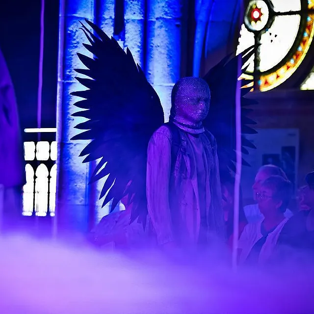 Eine dramatische Bühnenaufführung in einer Kirche unter intensivem blauem Licht, mit einer zentralen Figur mit großen schwarzen Flügeln und verschleiertem Gesicht, die inmitten von dichtem Bühnennebel steht.