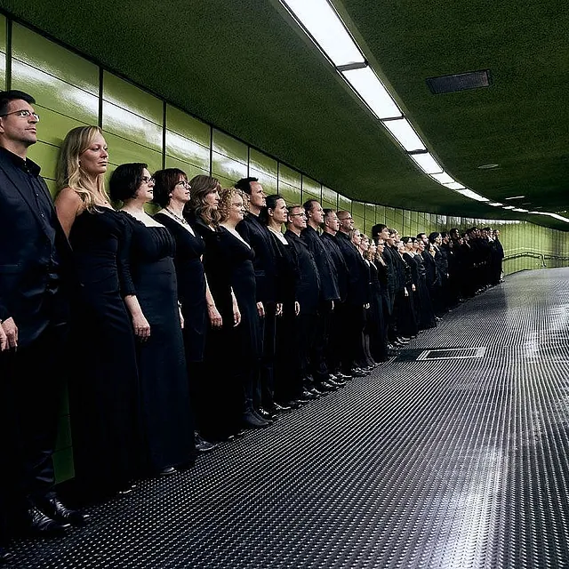 Ein großer Chor in formeller schwarzer Kleidung steht in einer langen, geordneten Reihe an der geschwungenen grünen Fliesenwand eines modernen Fußgängertunnels.
